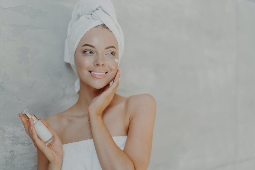 Pleased European woman puts on moisturizing cream on face, looks gladfully aside